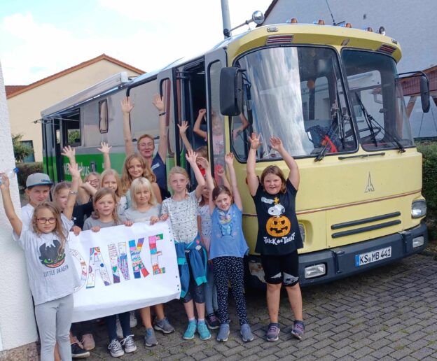 Das Bild zeigt eine glückliche Gruppe Mädchen vor dem Mädchenbus, um den es in diesem Vortrag geht. Die Mädchen halten ein Schild, auf dem in Großbuchstaben "Danke" steht.